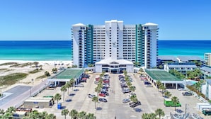A high-rise hotel with a central entrance, multiple floors, and a parking lot with cars and palm trees. - Seasational Escape!  Amazing Beachfront Condo/Covered Balcony, BEACH SER, Pool (Panama City Beach)