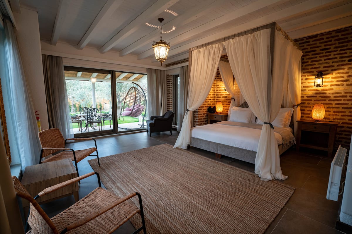 A spacious bedroom with a large bed, a wicker chair, a dining area with a table and chairs, and a view of the outdoors.