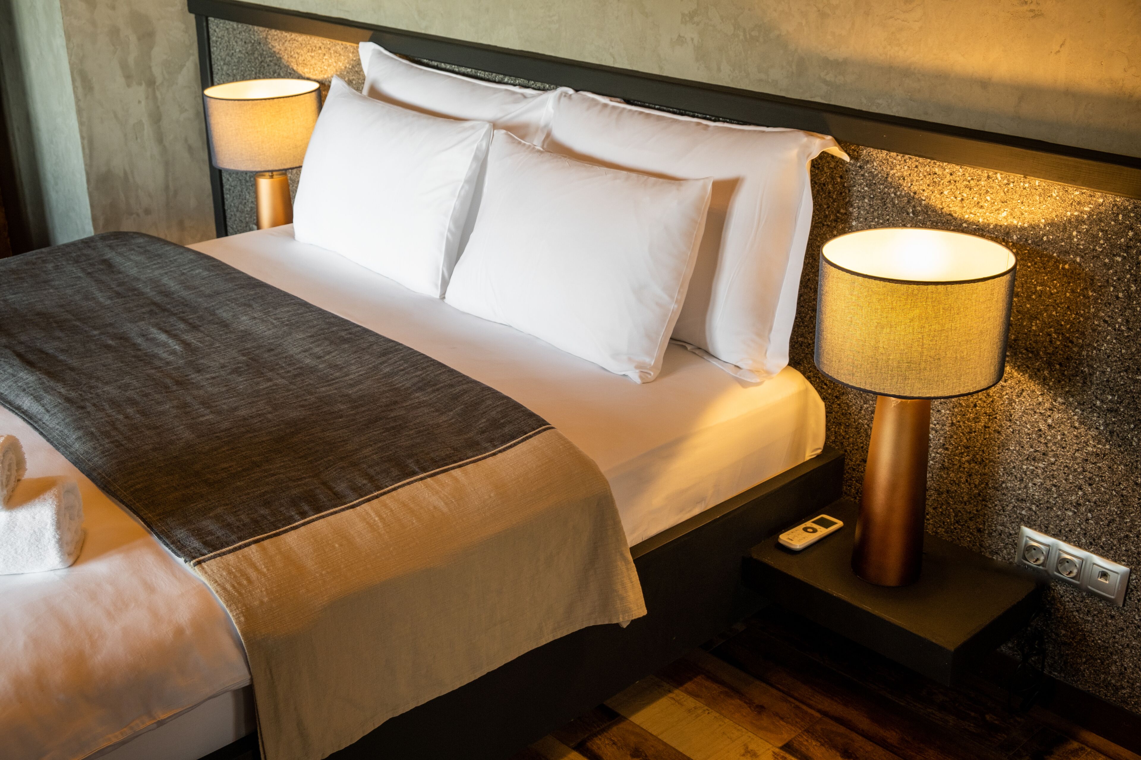 Een net opgemaakt bed met witte lakens en een grijze plaid, met aan beide zijden nachtkastjes met bijpassende bedlampen.