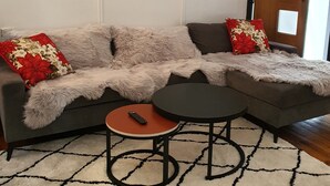 A living room with a grey sofa, two red and white floral cushions, a round coffee table, and a framed picture on the wall. - Rustic large 5 bedroom home in a inner city suburb (Enfield)