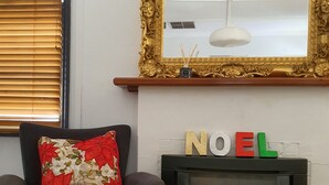 A living room with a fireplace, a sofa with a red floral pillow, and a mirror with a golden frame above it. - Rustic large 5 bedroom home in a inner city suburb (Enfield)