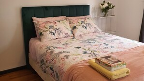 A neatly made bed with a floral bedspread, a green upholstered headboard, and a small bedside table with a vase of flowers. - Rustic large 5 bedroom home in a inner city suburb (Enfield)