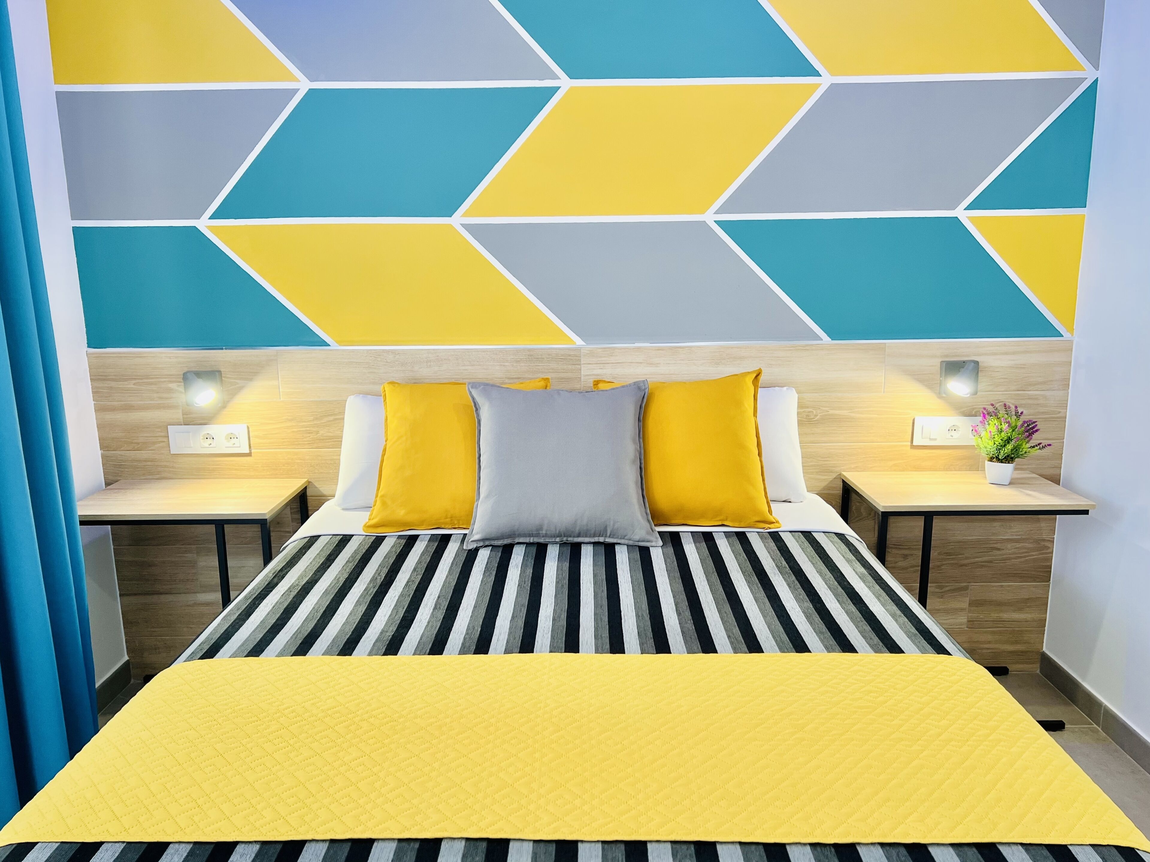 Quarto de hotel com uma cama, uma mesa de cabeceira iluminada por um abajur e uma parede com padrões geométricos.
