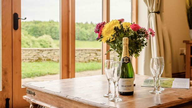 A wooden table with a bottle of Delacourt champagne, two glasses, and a bouquet of flowers. - Dog friendly holiday cottage in the Cotswolds with a hot tub - Owls Roost (Cold Aston, near Bourton-on-the-Water)