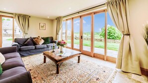 A spacious living room with a sofa, a coffee table, and a view of a green landscape through large sliding glass doors. - Dog friendly holiday cottage in the Cotswolds with a hot tub - Owls Roost (Cold Aston, near Bourton-on-the-Water)