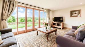 A living room with a large sliding glass door leading to a garden, a wooden coffee table, a sofa, a TV on a wooden stand, and a framed picture on the wall. - Dog friendly holiday cottage in the Cotswolds with a hot tub - Owls Roost (Cold Aston, near Bourton-on-the-Water)