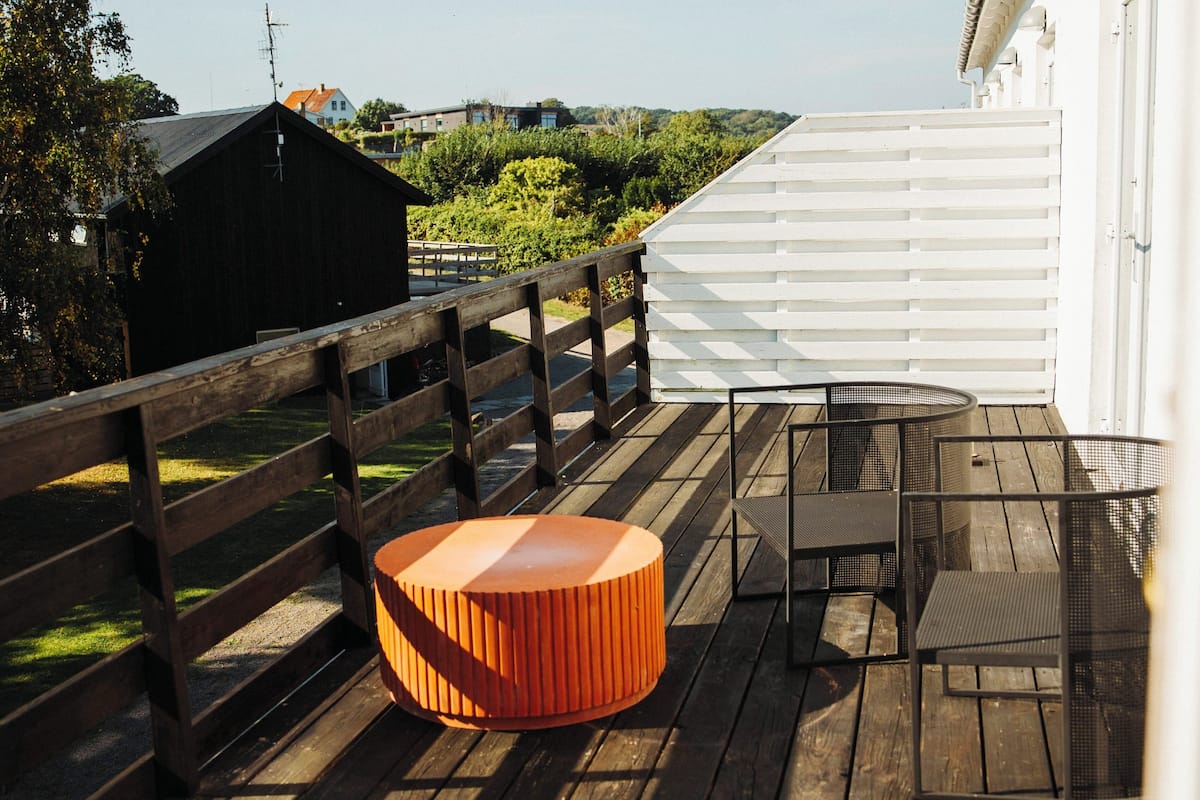 Una terraza de madera con un otomano naranja redondo, sillas de metal y una valla blanca.