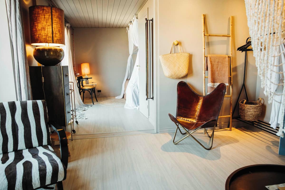 Una habitación con una silla a rayas, una silla de cuero, una lámpara, una escalera y una cesta tejida.