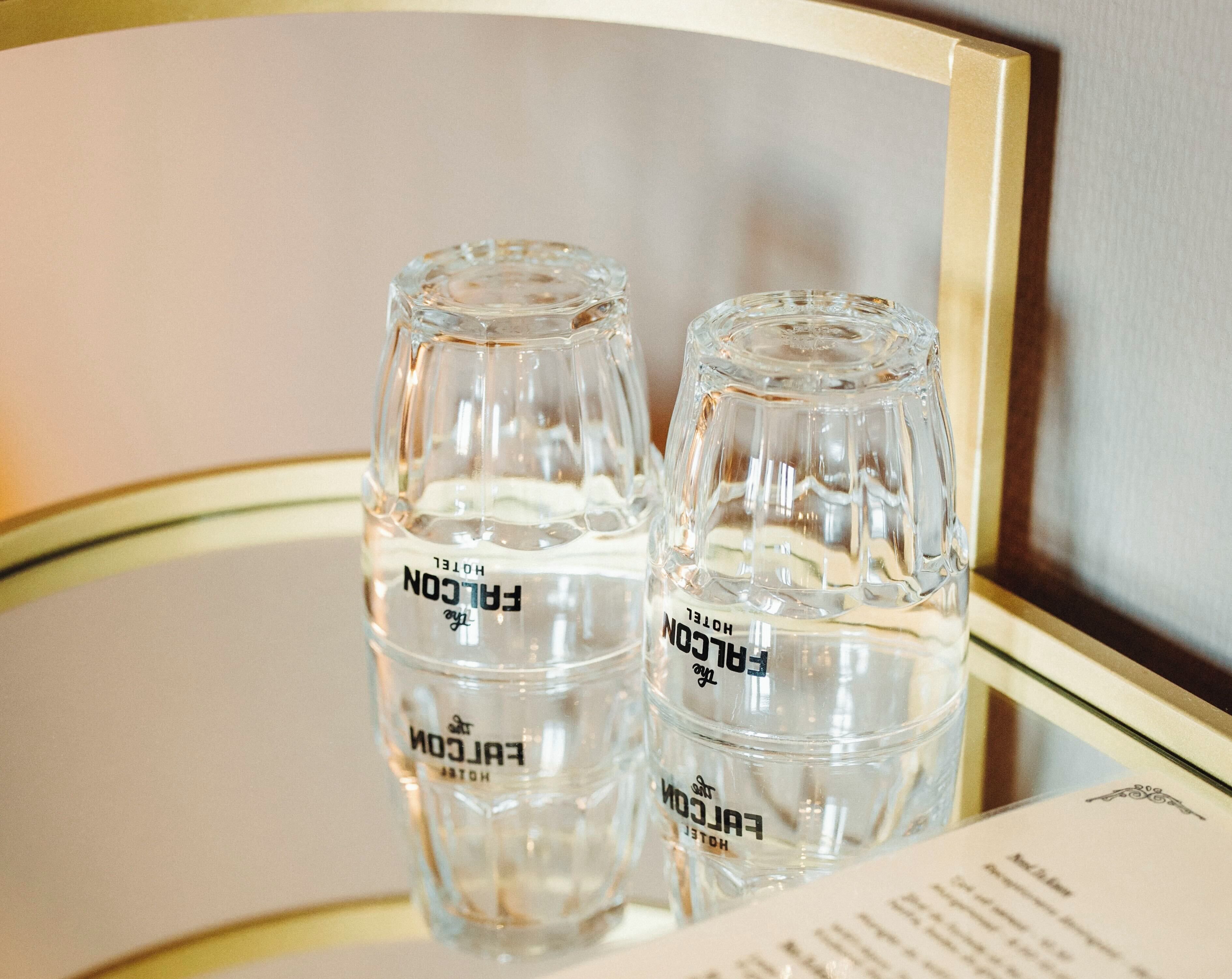Deux bouteilles en verre transparent portant l’étiquette « FALCON HOTEL », posées sur une surface réfléchissante, avec une partie d’un menu ou d’un dépliant visible.