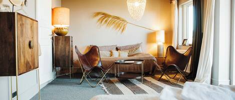 A hotel room with a bed, two chairs, a small table, a lamp, a hanging light fixture, and a window with curtains.