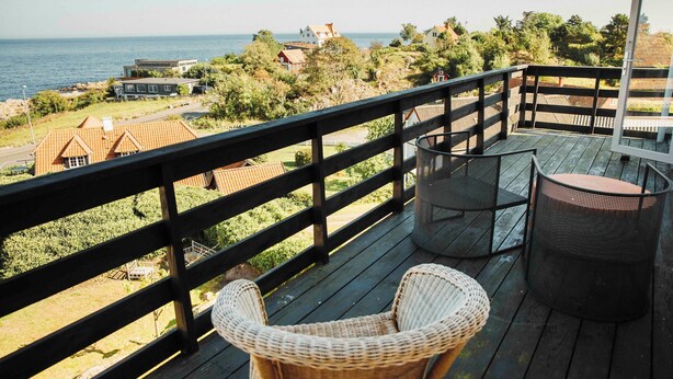 Une terrasse en bois, agrémentée de chaises et d’une table en osier, qui offre une vue sur une zone côtière avec des maisons et de la végétation.