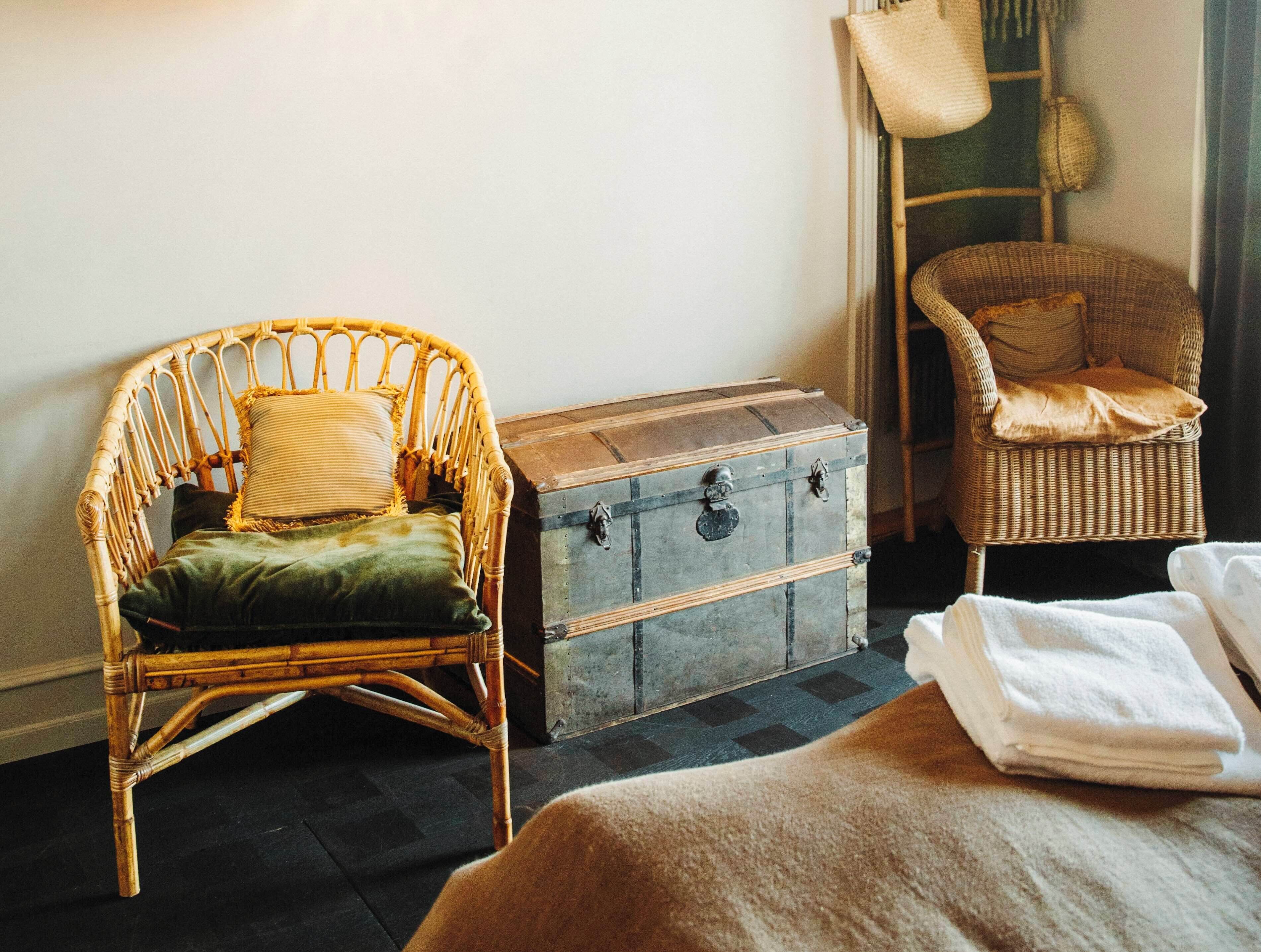 A room with a wicker chair, a wooden chest, and a bed with white linens.