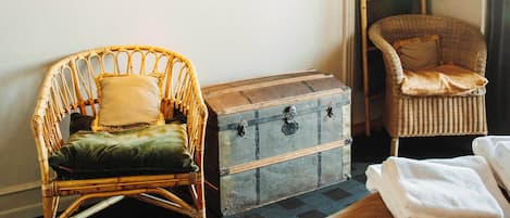 A room with a wicker chair, a wooden chest, and a bed with white linens.