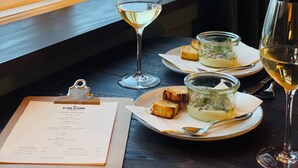Une table avec un menu, deux verres de vin blanc, un petit pot de nourriture et une assiette de pain, le tout placé près d’une fenêtre offrant une belle vue.