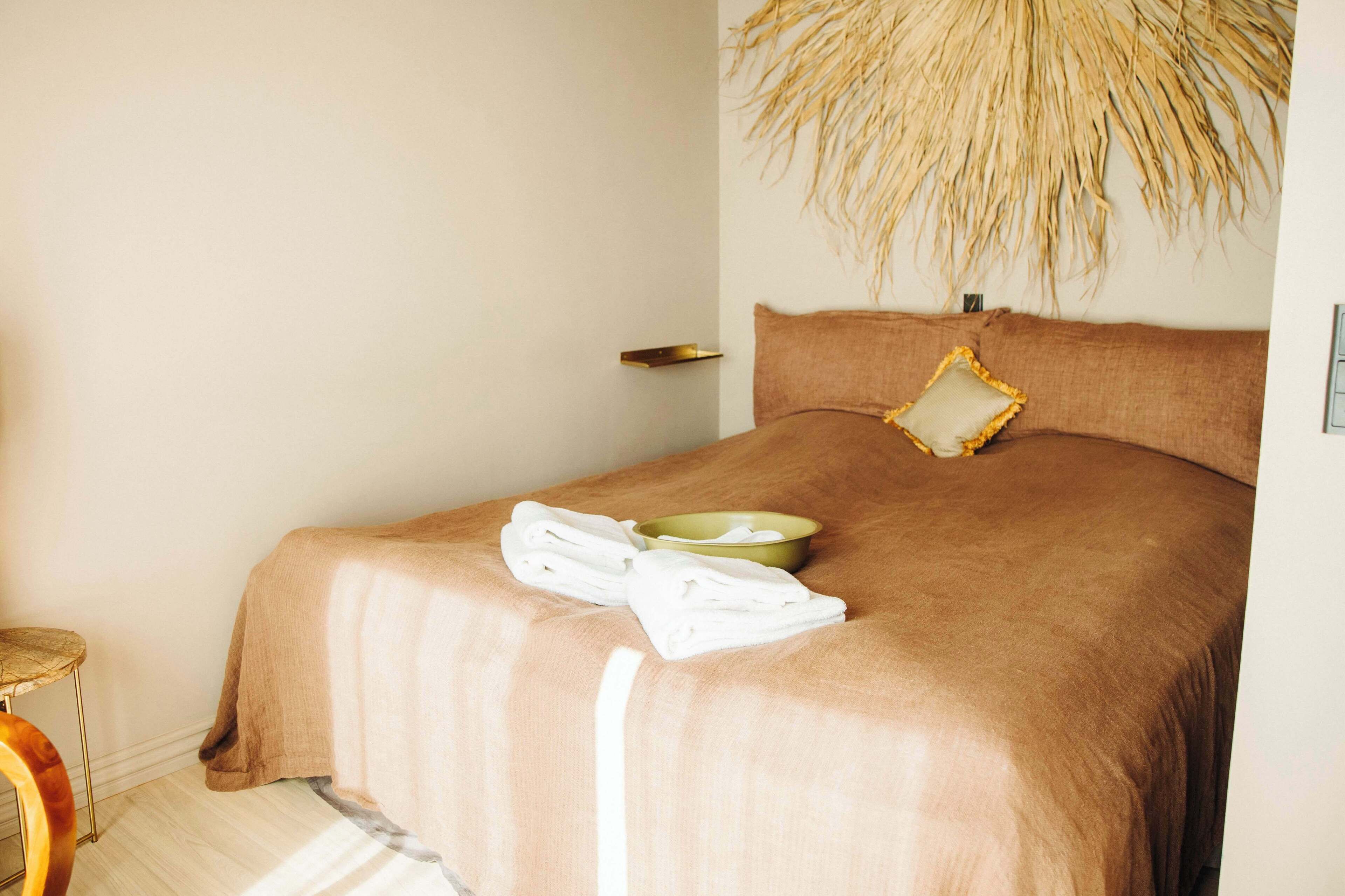 A neatly made bed with a brown bedspread and a single pillow, a green bowl, and a pair of folded white towels on the bed.