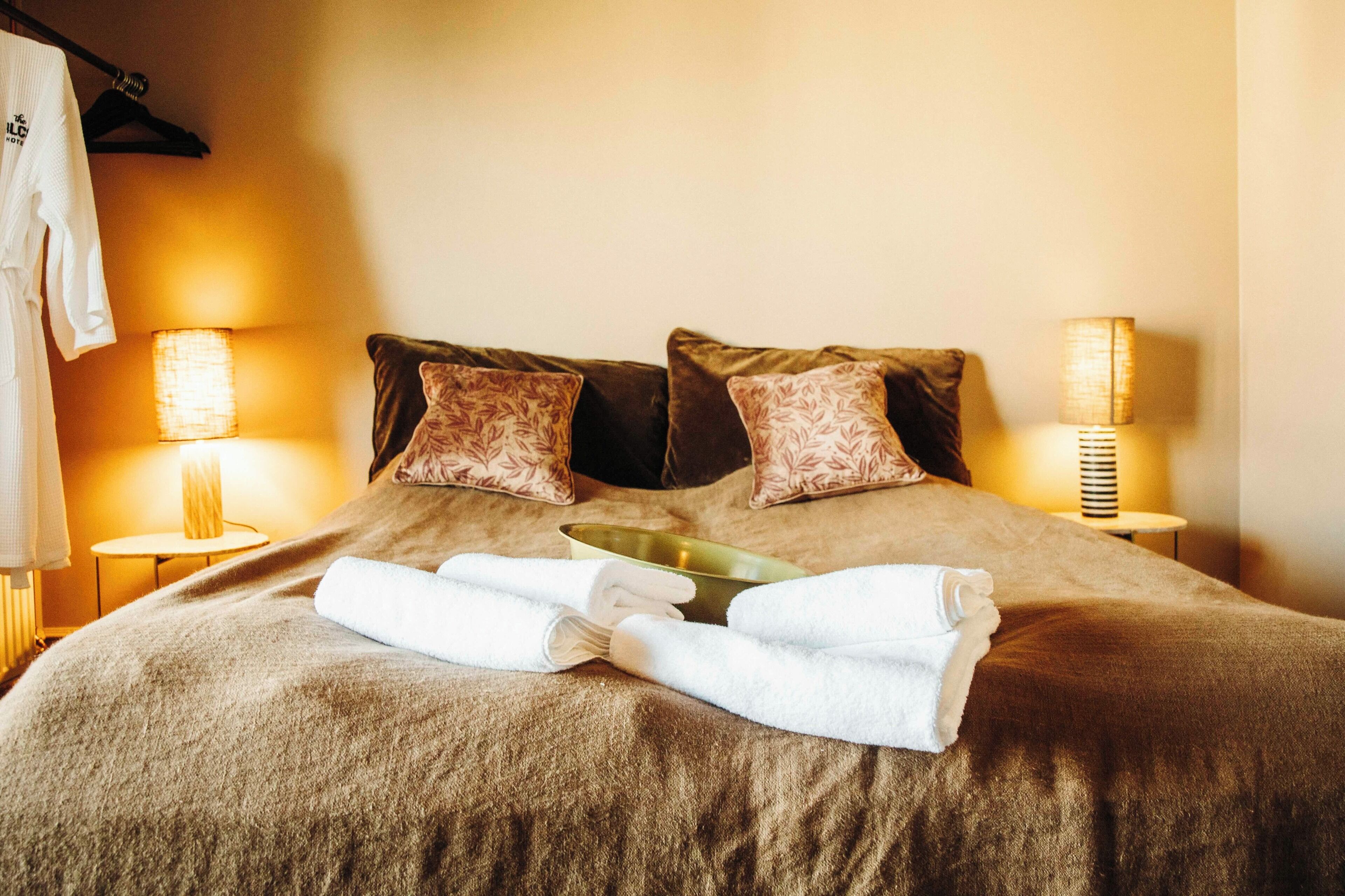 A neatly made bed with a brown blanket, two decorative pillows, and a set of folded white towels. There are two bedside lamps with lampshades and a hanging robe on a rack.