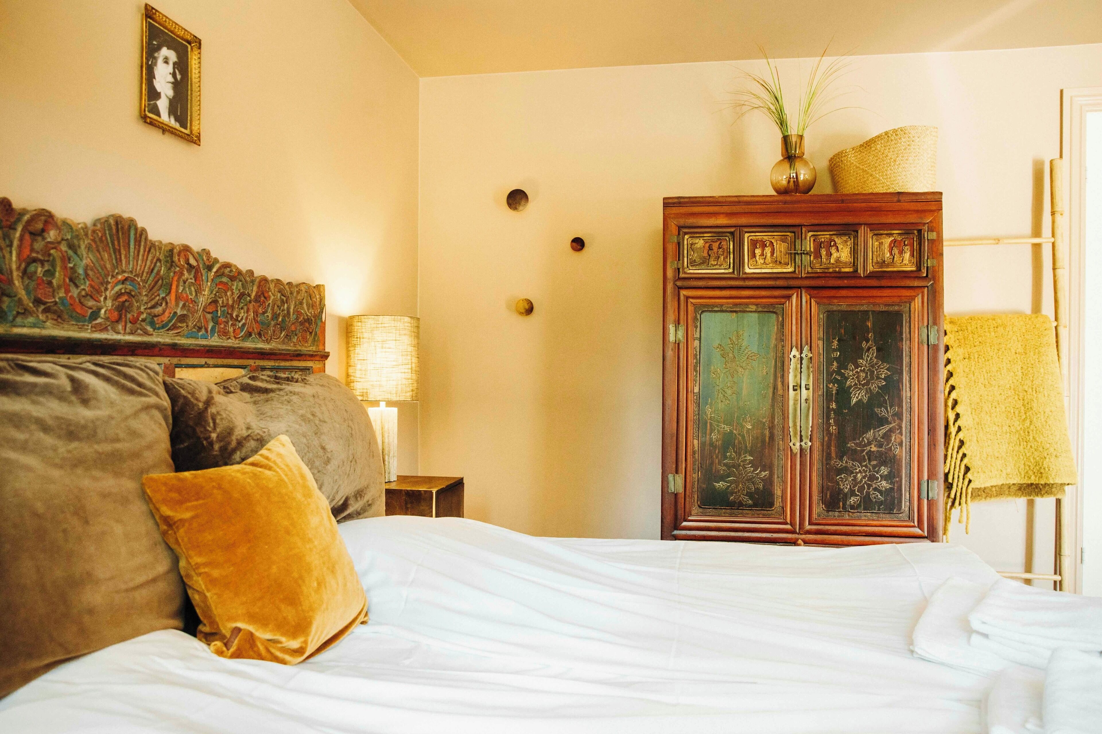 Een opgemaakt bed met hoofdbord, een houten kastje met decoratieve panelen en een lamp op een nachtkastje.