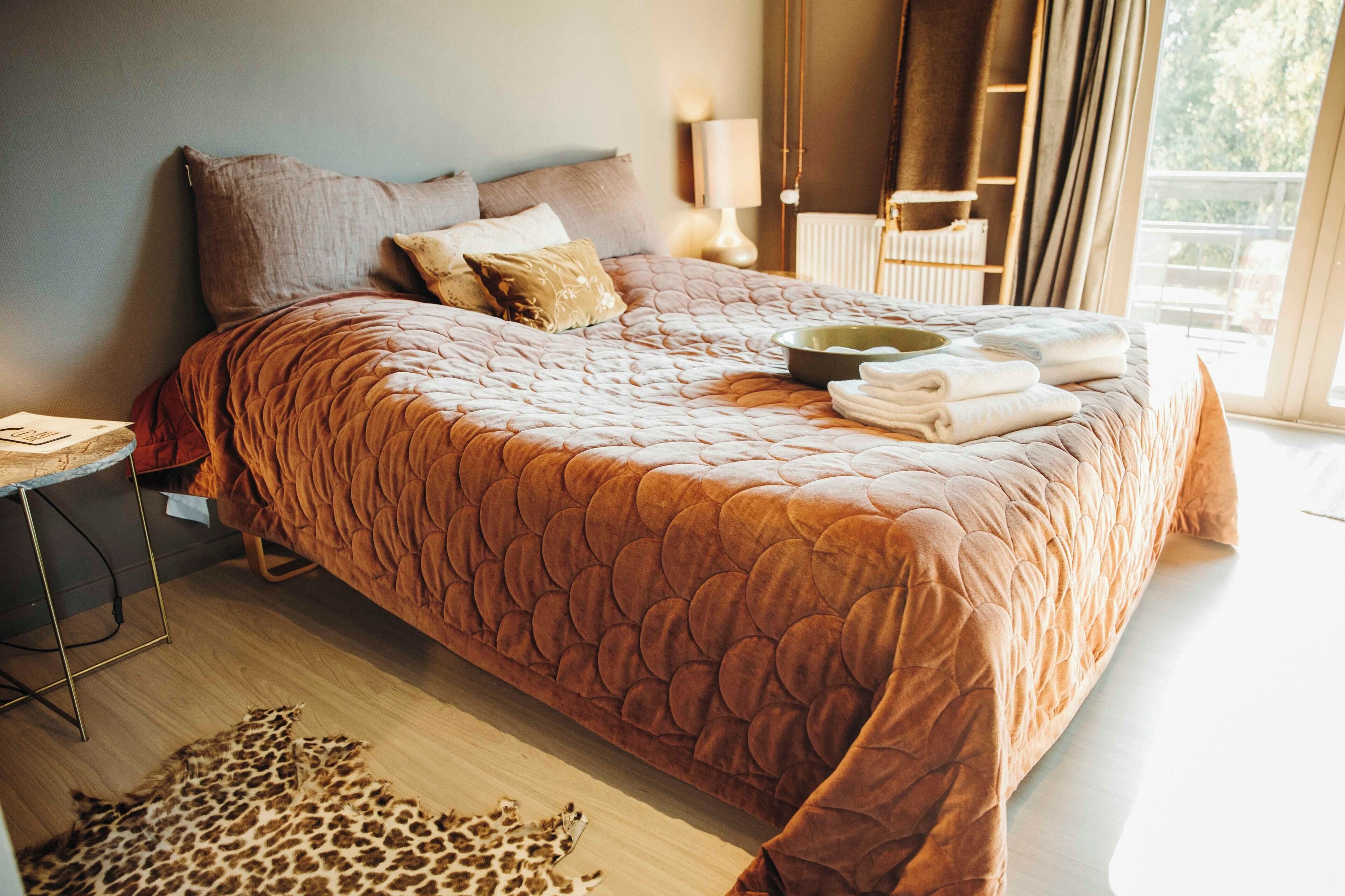 A large bed with a brown quilt, pillows, and a bowl of towels.