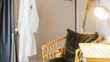 A room with a bed, a chair with a green cushion, a table, and a clothing rack with white robes.