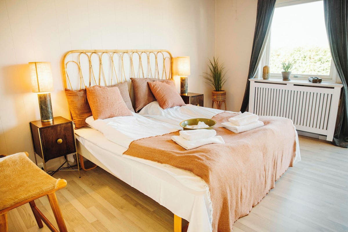 Una cama bien hecha con cabecera, dos mesitas de noche con lámparas, una planta en maceta y una ventana con cortinas.