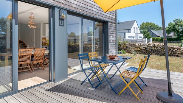 A modern house with a wooden deck, a dining area with a table and chairs, and a large yellow umbrella.