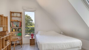A bedroom with a sloped ceiling, a white bed, a wooden bookshelf, a chair, and a window with a view of greenery.