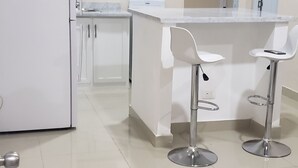 A modern kitchen with white cabinets, a white refrigerator, a white countertop, and two white bar stools.