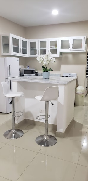 Uma cozinha moderna com armários brancos, uma ilha central com dois bancos altos, uma geladeira, um micro-ondas e um fogão a gás.