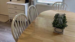 Uma sala de jantar com mesa de madeira, cadeiras brancas, uma planta em vaso e vista para o verde lá fora.