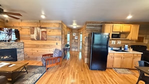 A spacious living area with a fireplace, wooden flooring, and a modern kitchen with wooden cabinets and appliances. - Chimney Mtn 1 -  2BR Cabin, Dog Friendly, Fireplace, Covered Hot Tub. Near Helen (Sautee Nacoochee)