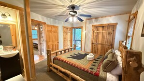 A room with a bed, a ceiling fan, wooden doors, and a bathroom with a mirror and sink. - Chimney Mtn 1 -  2BR Cabin, Dog Friendly, Fireplace, Covered Hot Tub. Near Helen (Sautee Nacoochee)