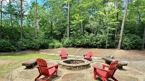 A circular outdoor seating area with a fire pit, surrounded by red Adirondack chairs, set against a backdrop of trees. - Chimney Mtn 1 -  2BR Cabin, Dog Friendly, Fireplace, Covered Hot Tub. Near Helen (Sautee Nacoochee)