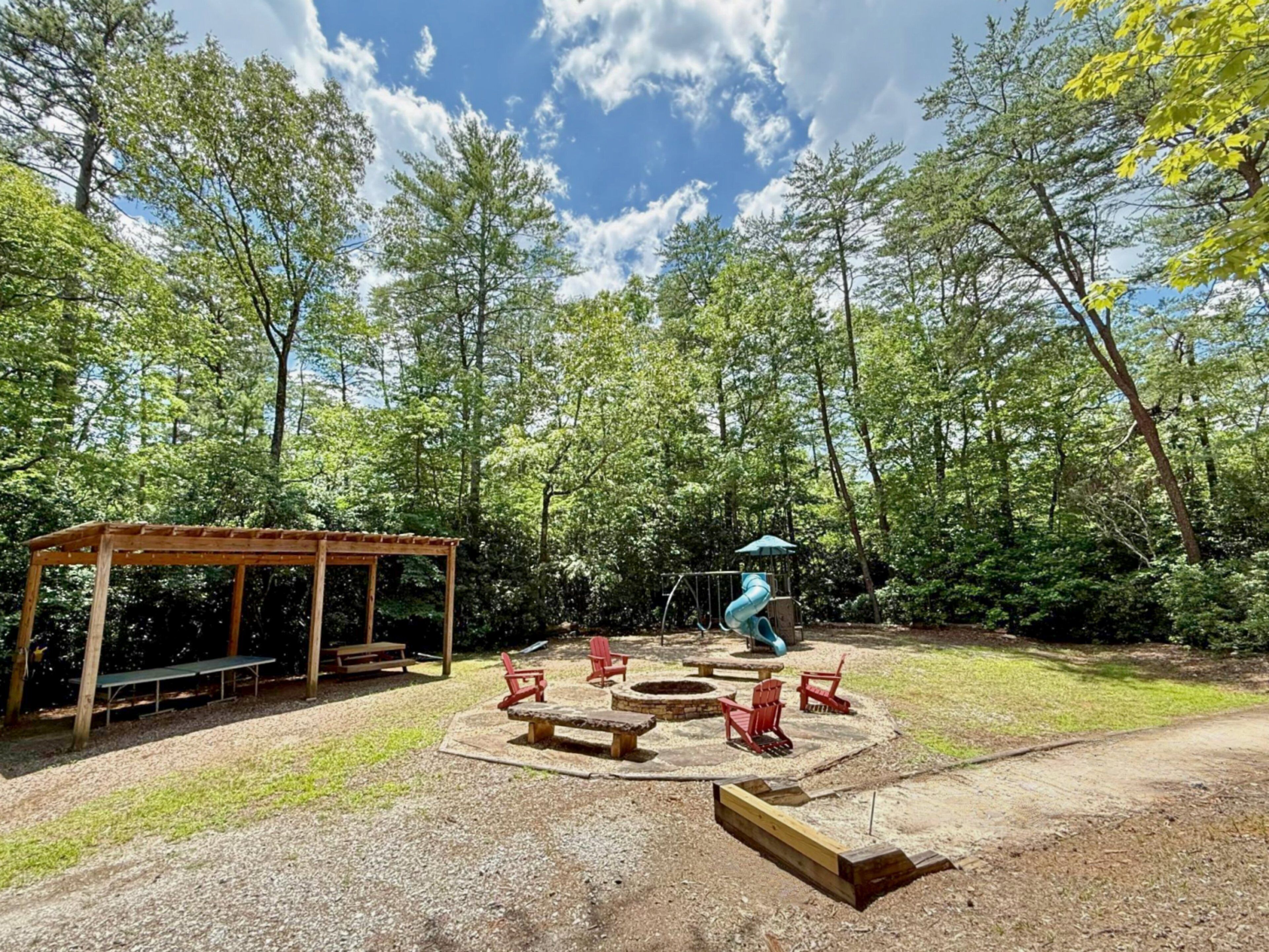 Metsäinen ulkoalue, jossa on puinen huvimaja, nuotiopaikka ja punaiset Adirondack-tyyliset puutarhatuolit.