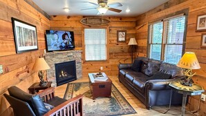 L’intérieur chaleureux d’un chalet avec une cheminée, un canapé en cuir, des murs en bois et un ventilateur de plafond.