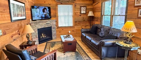 A cozy cabin interior with a fireplace, leather sofa, wooden walls, and a ceiling fan.