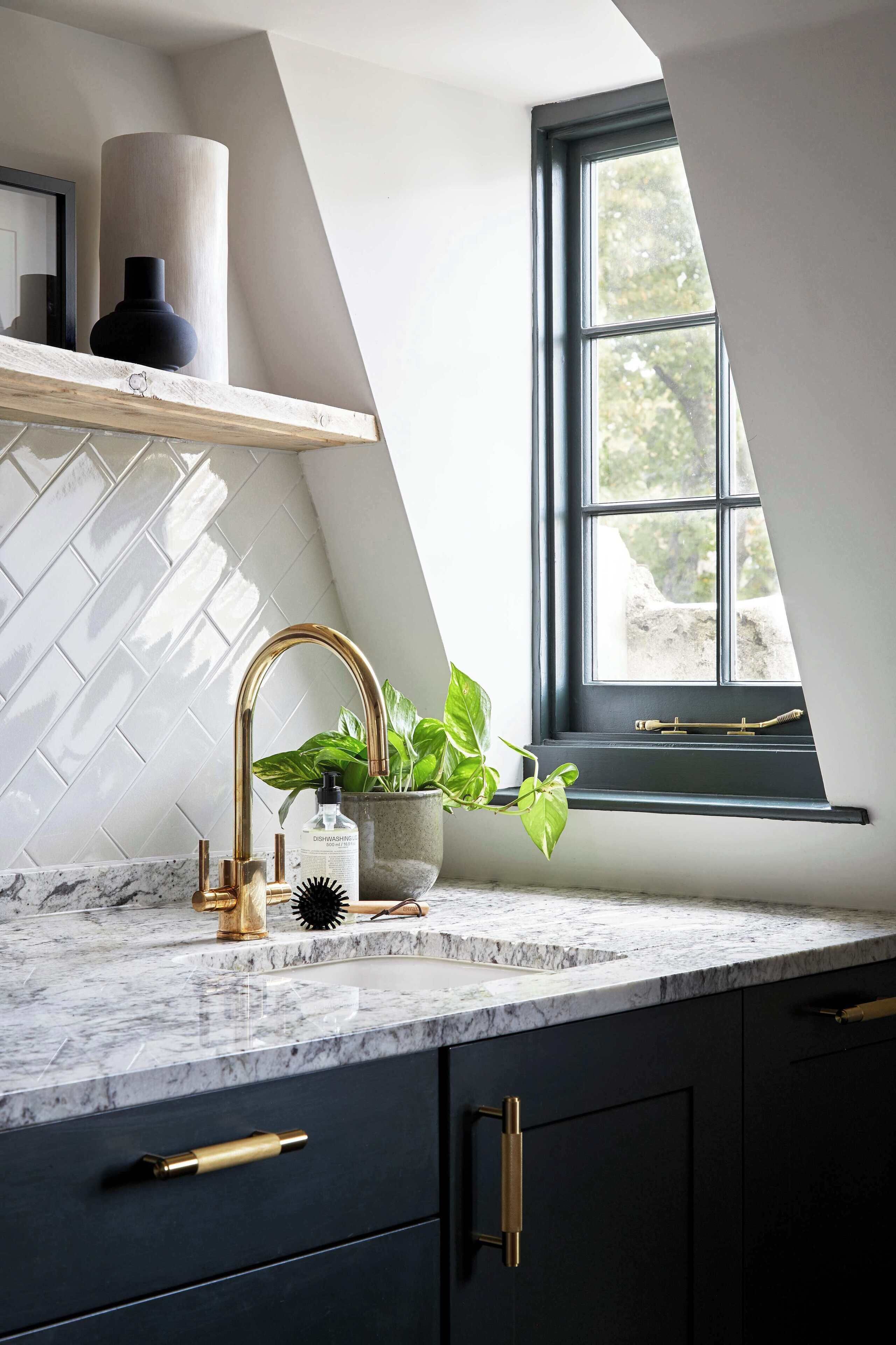 A modern kitchen with a marble countertop, a gold faucet, and a window with a view of greenery.