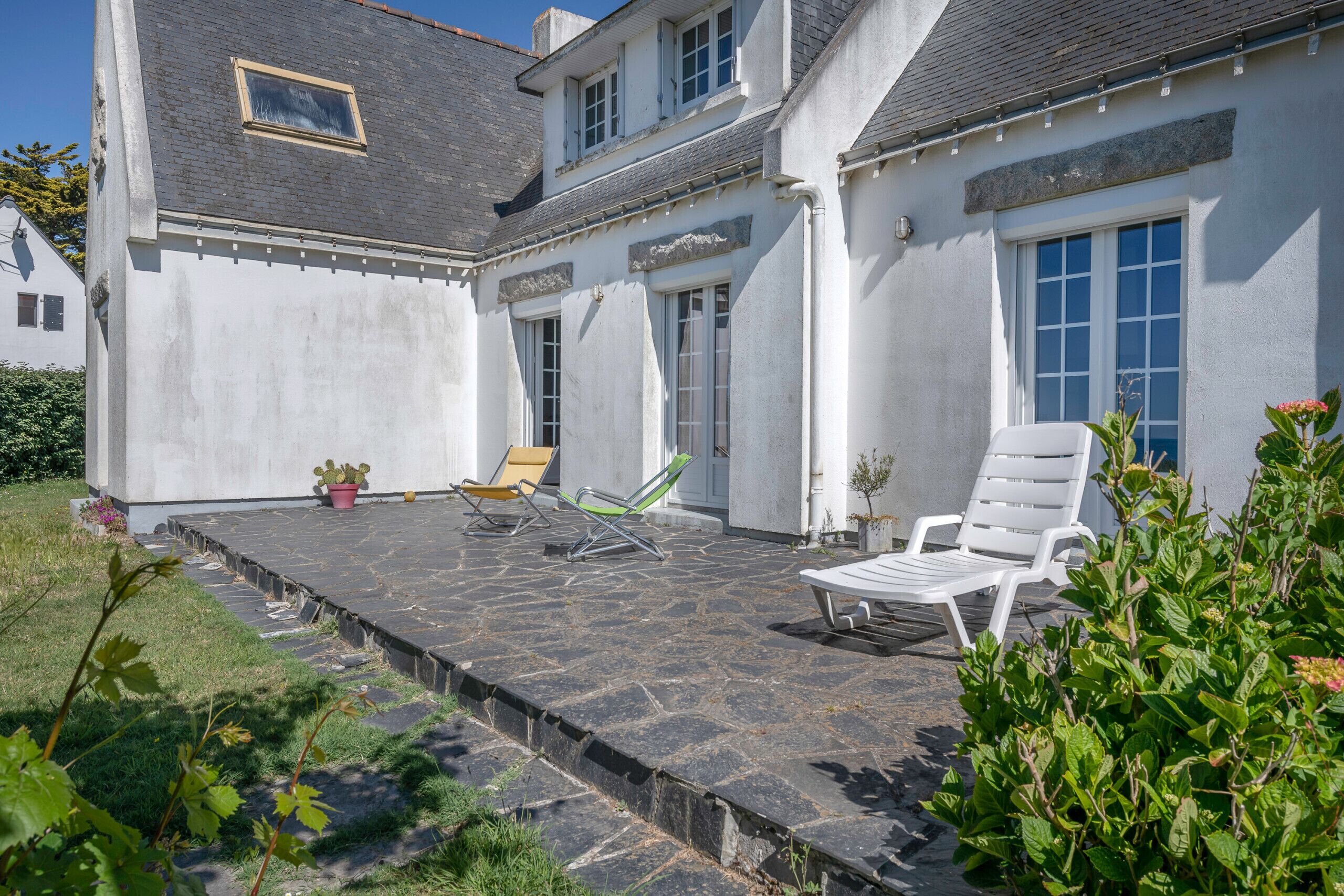 Uma casa com um alpendre de pedra, fachada branca e área de jardim com uma espreguiçadeira branca e uma cadeira amarela.
