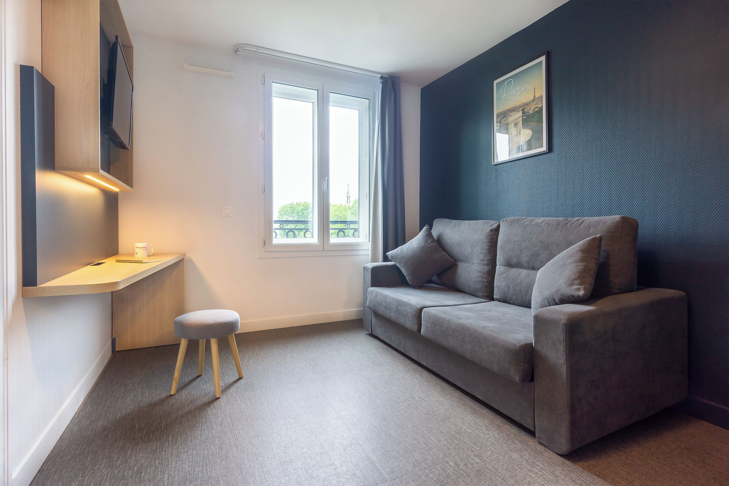 A modern room with a grey sofa, a small wooden stool, a desk with a lamp, and a large window with a view of a building.