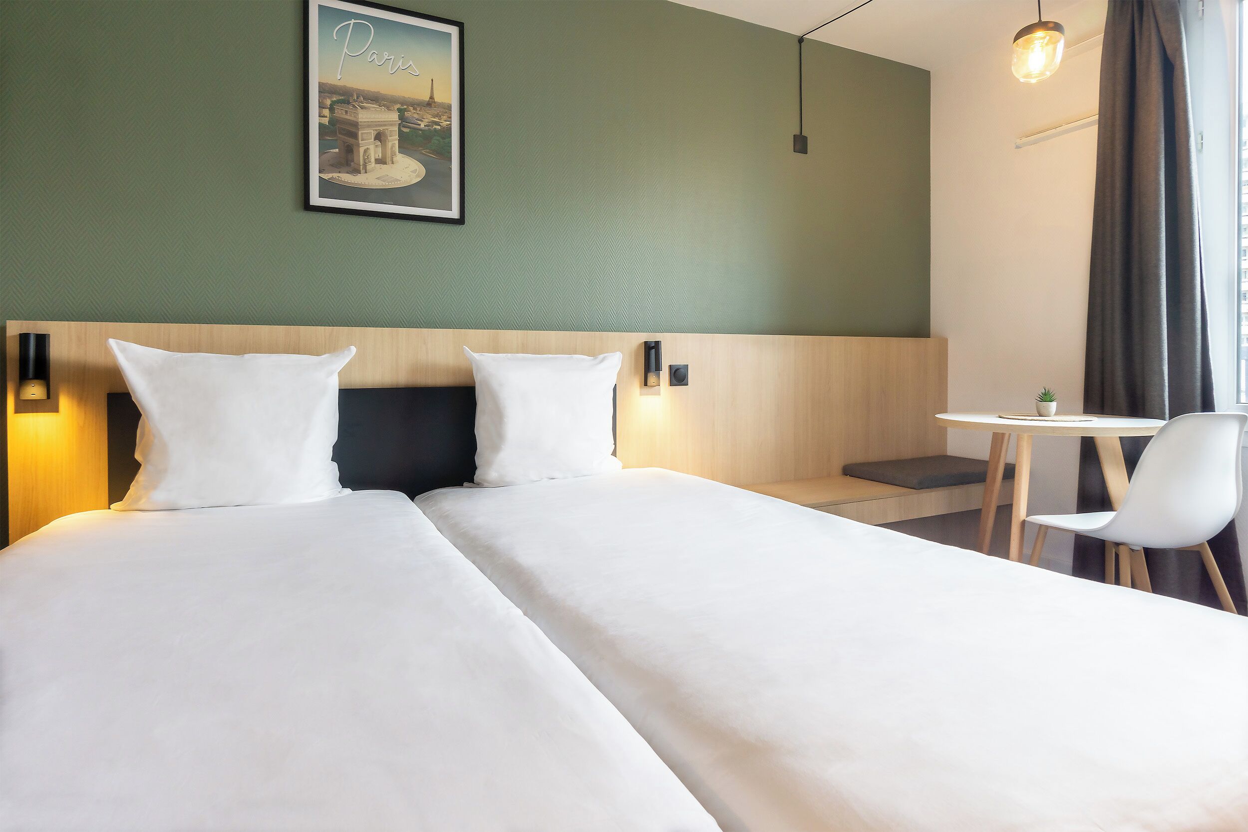 A hotel room with a bed, two bedside tables, a small table with a chair, a wall-mounted light, and a framed picture on the wall.
