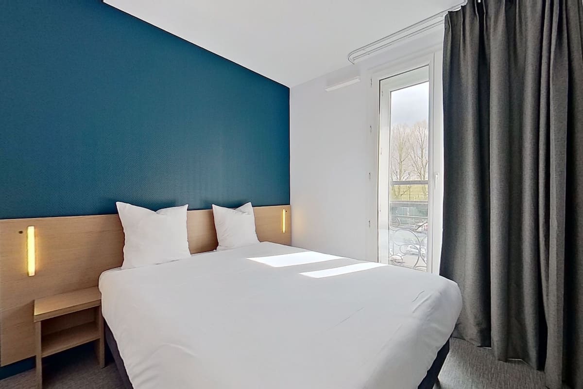 A hotel room with a large bed, a wooden nightstand, a blue accent wall, and a window with curtains.