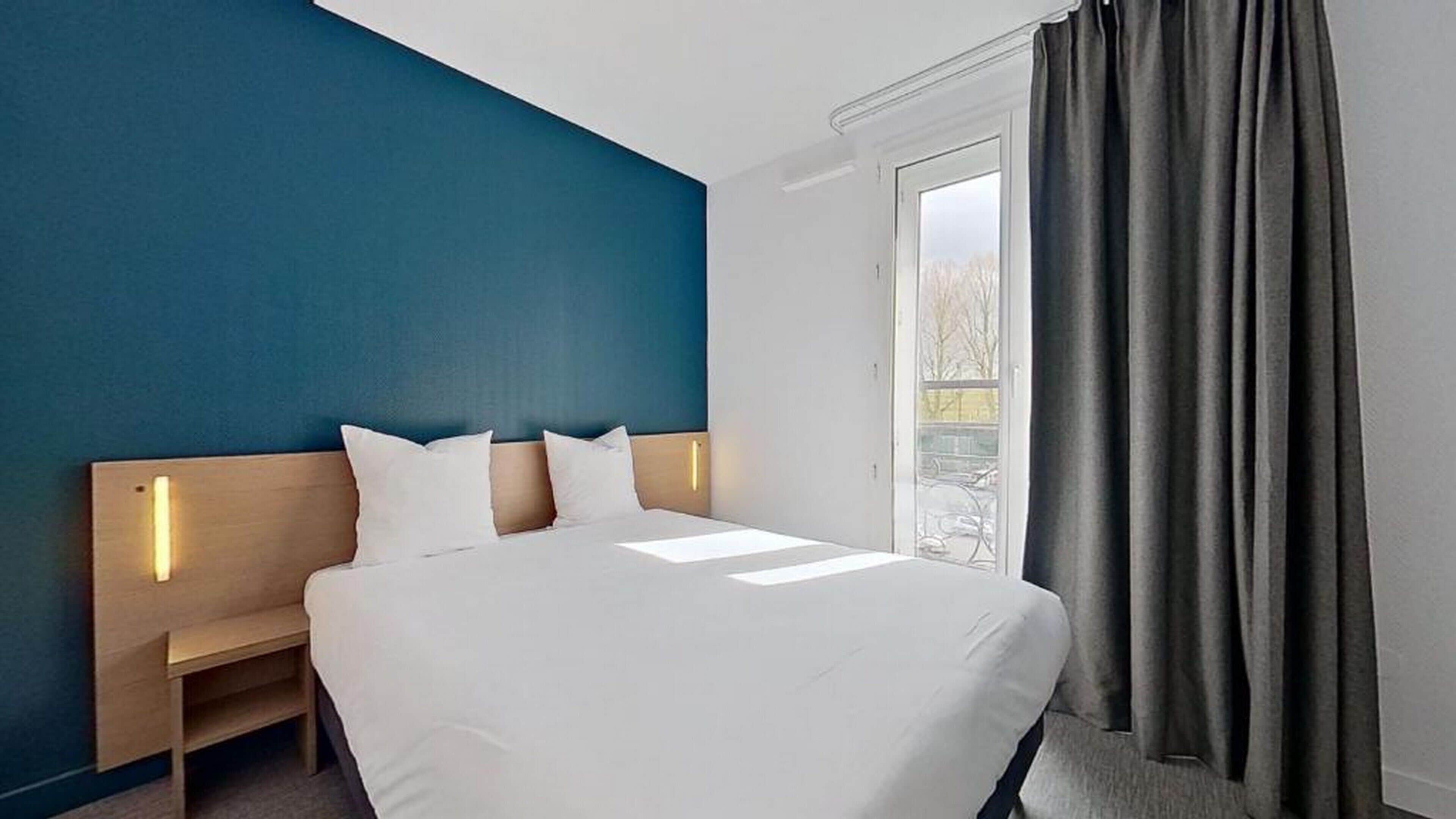 Une chambre d’hôtel avec un lit, une table de chevet, une fenêtre avec des rideaux et une vue sur les arbres à l’extérieur.