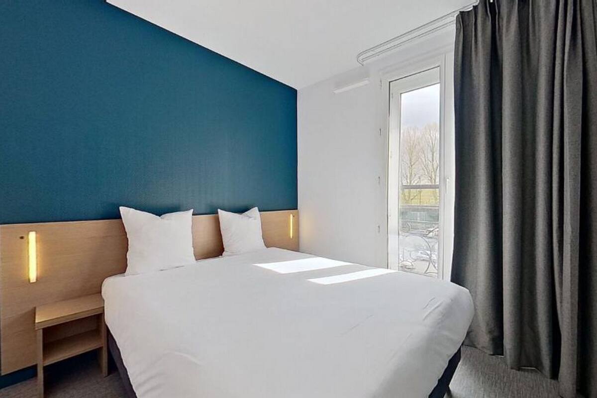 A hotel room with a bed, a nightstand, a window with curtains, and a view of trees outside.