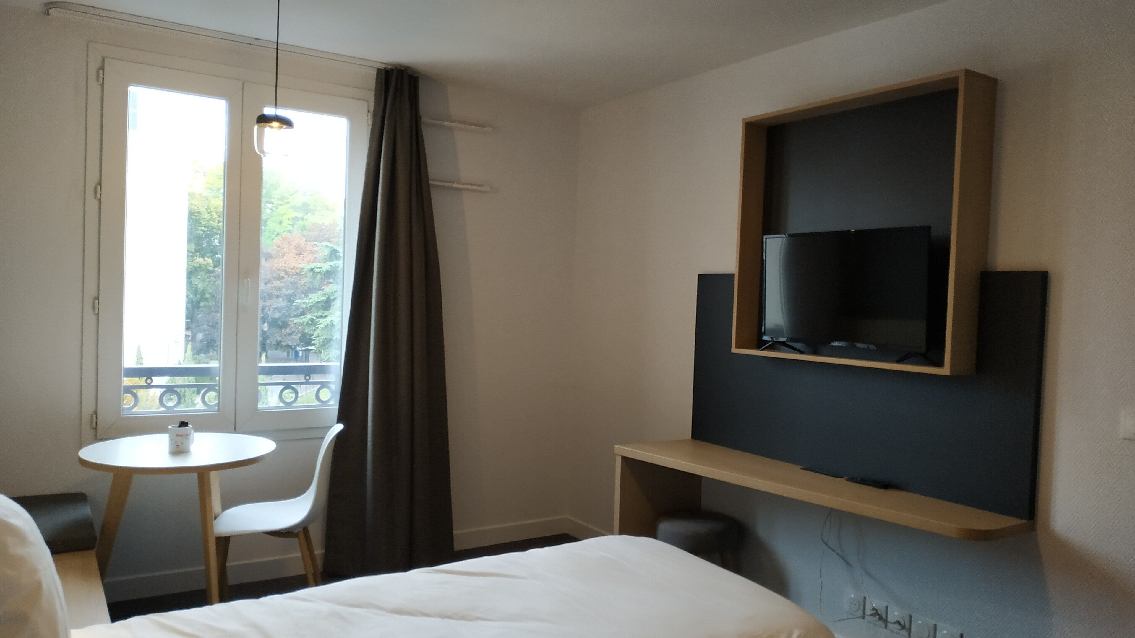 A hotel room with a bed, a TV mounted on the wall, a small table with a chair, and a window with curtains.