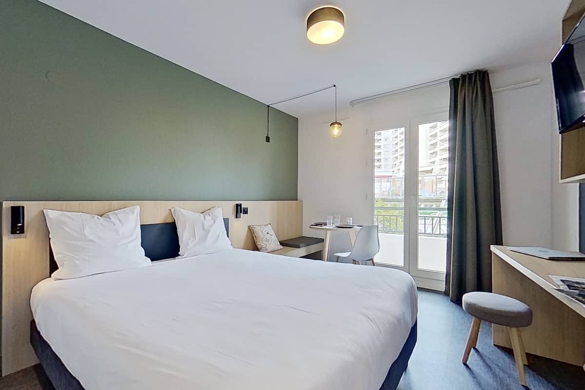 A hotel room with a large bed, a desk, a chair, a TV mounted on the wall, and a window with curtains.