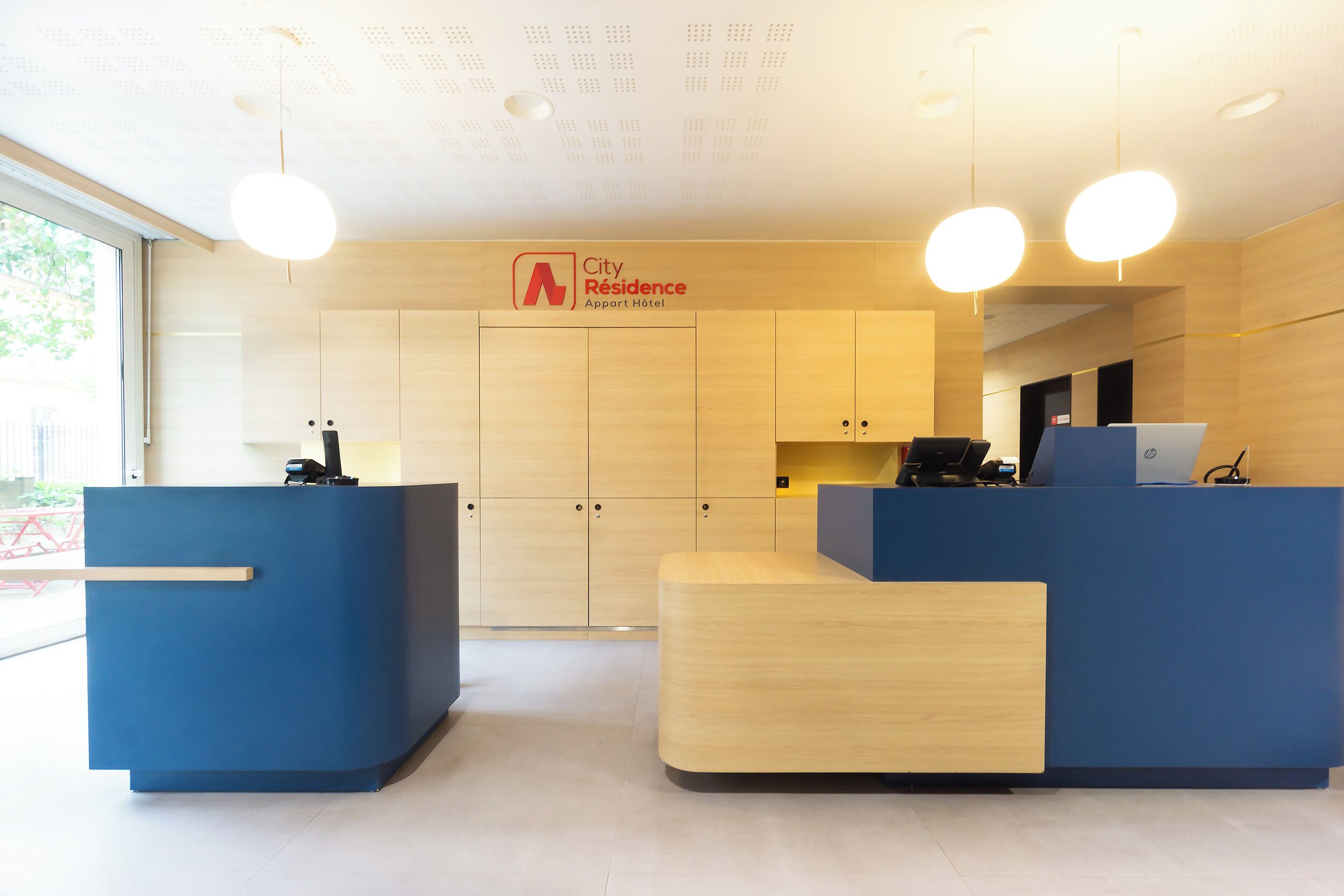 A hotel reception area with a modern design, featuring a blue reception desk, a wooden counter, and a sign that reads "City Residence Apartment Hotel".
