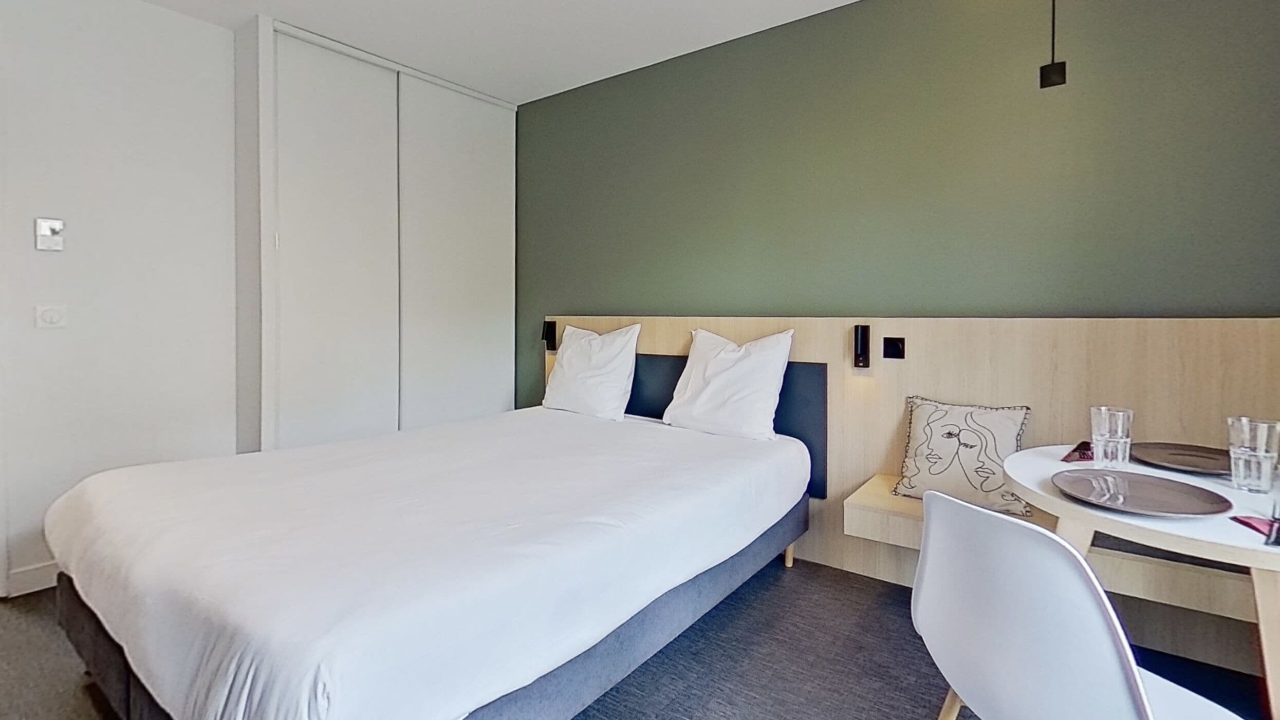 A modern hotel room with a large bed, a desk with a chair, and a small table with glasses and a plate.