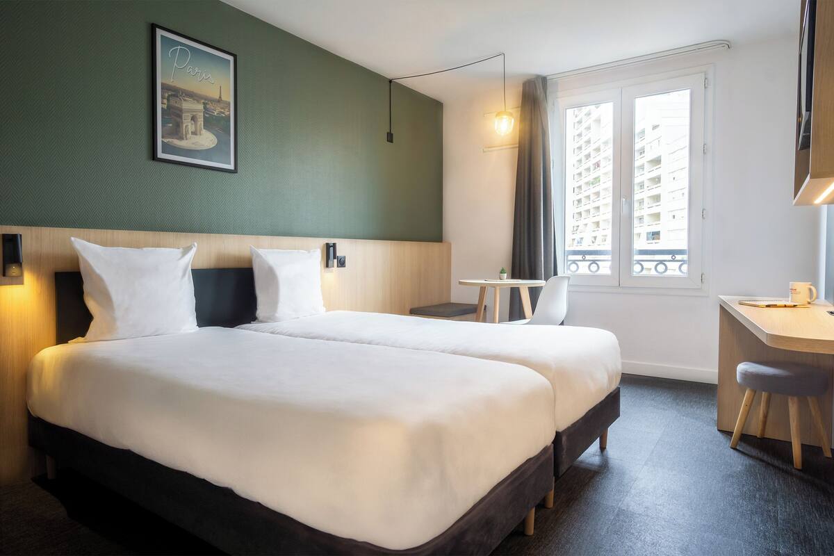 A hotel room with two beds, a desk, a chair, a small table, a lamp, and a window with a view of buildings.