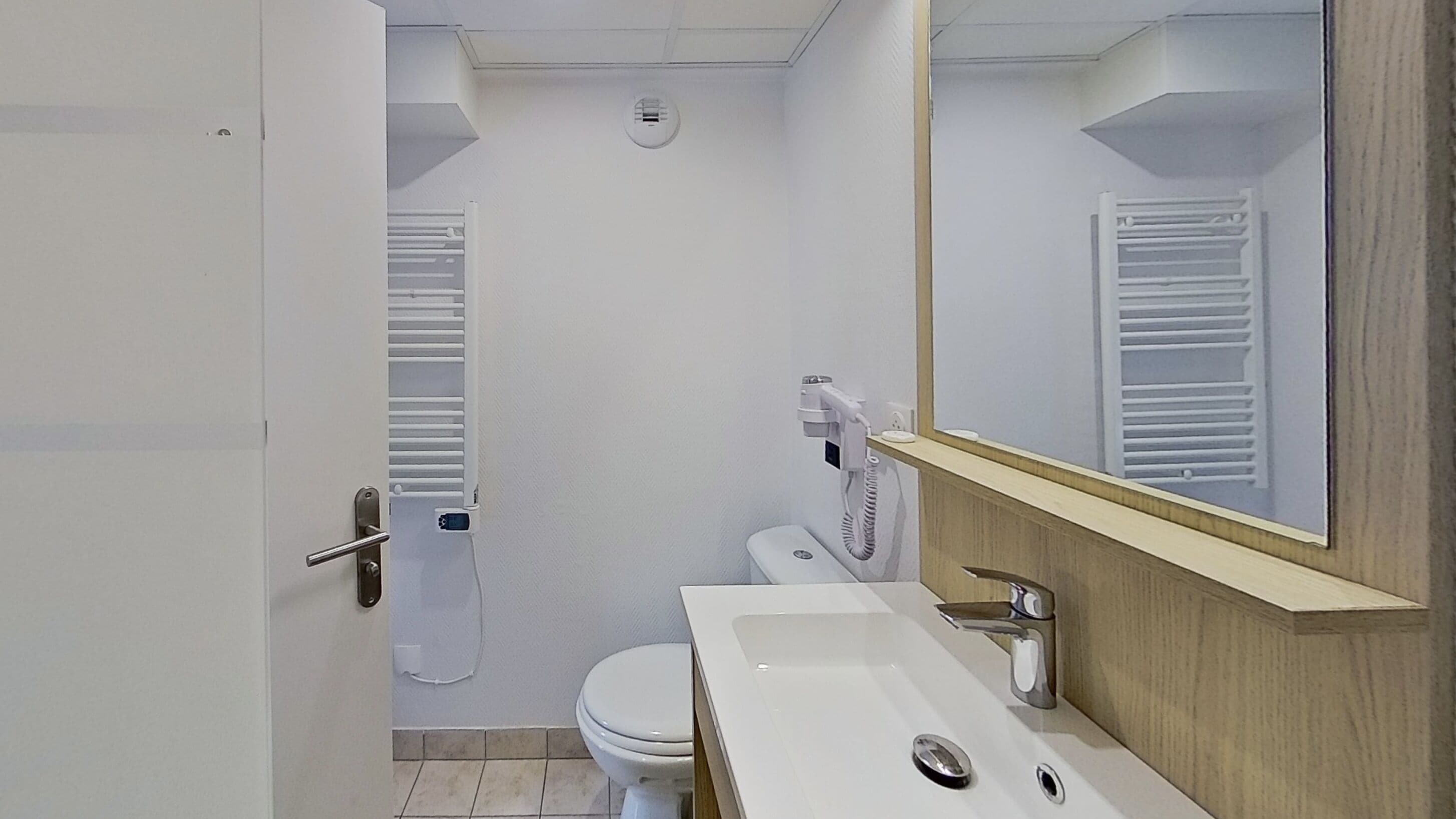 Et lille, rent badeværelse med et hvidt toilet, håndvask og et spejl over den. Der er en håndklædeholder på væggen og en varmeovn på gulvet.