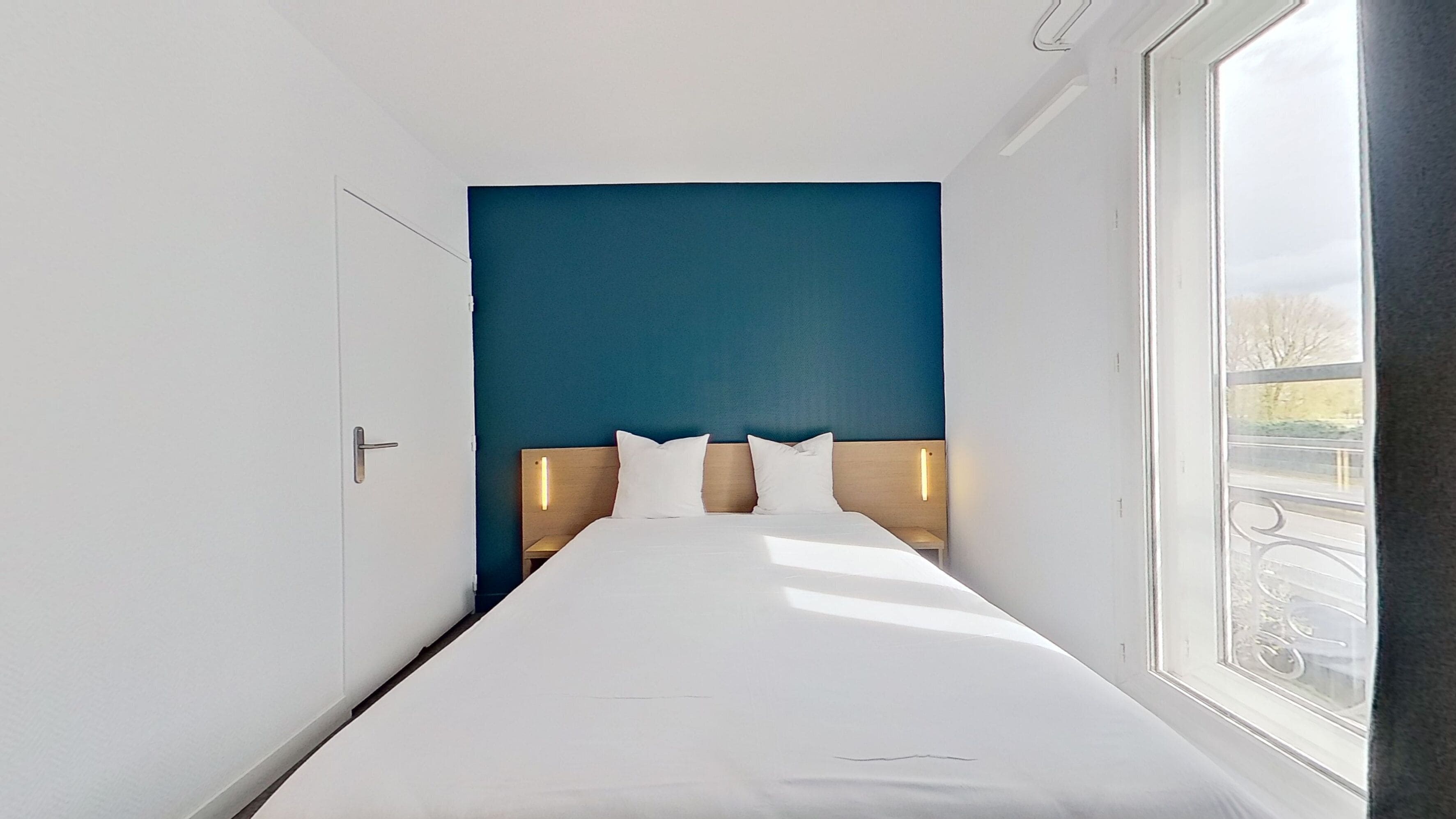 A modern hotel room with a large bed, a blue accent wall, and a view of the outdoors.