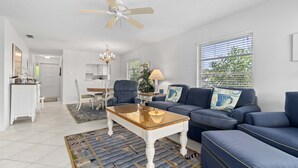 Un salon avec un canapé bleu, une table basse et un lustre. On y trouve également un ventilateur de plafond, une fenêtre avec des stores et une porte qui donne sur une autre pièce.